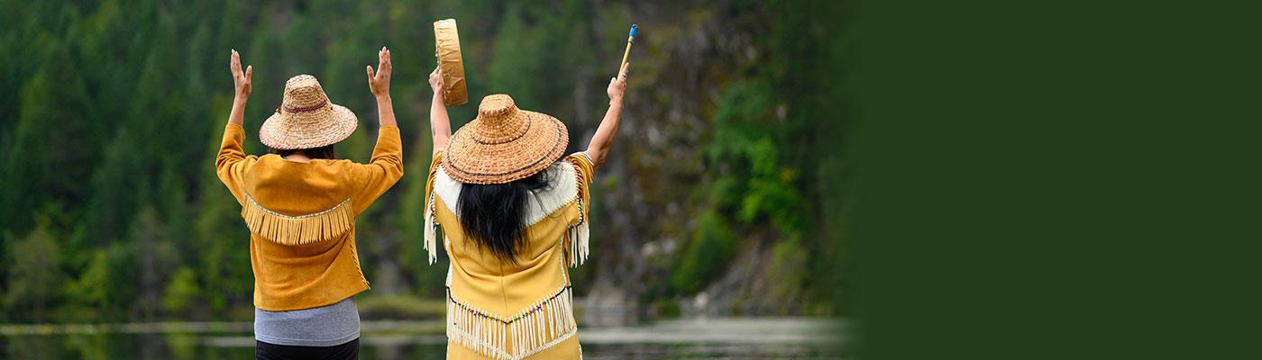 Two Native American women facing the forest, with their arms raised in dance.