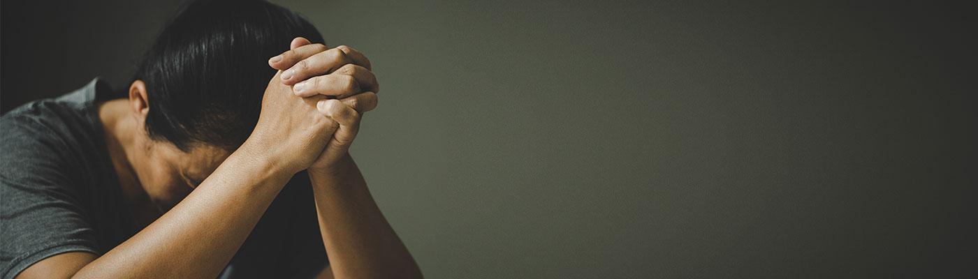 Young man with his elbows on a table and hands clasped, hangs his head and rests it on his hands..